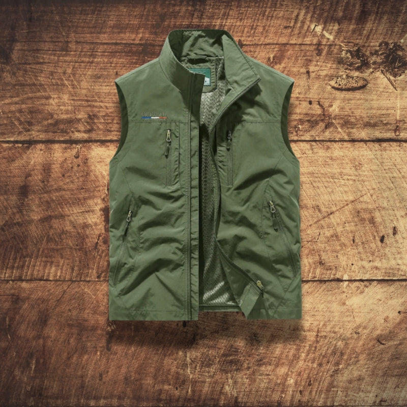 An olive green lightweight zip-up vest laid flat on a rustic wooden plank background. The vest features a high collar, a central zipper, zippered pockets, and a small colorful logo on the left chest.