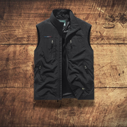 A black sleeveless zip-front vest with a stand collar and multiple zip pockets, laid flat on a rustic brown wooden plank background.