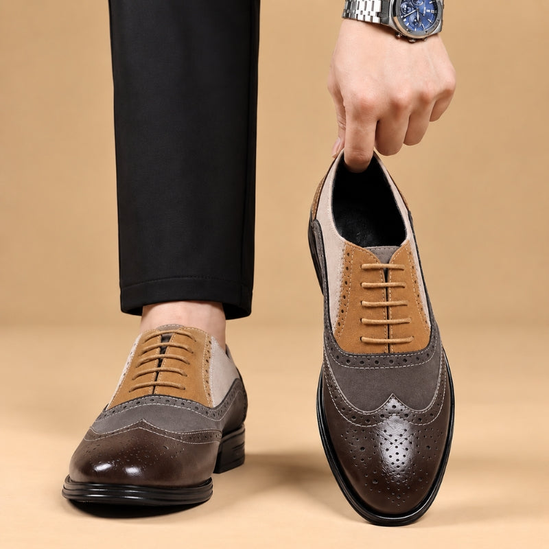 A pair of men's wingtip oxford shoes in brown, tan, and gray tones, shown on a model's feet and held by a hand against a warm beige studio background.