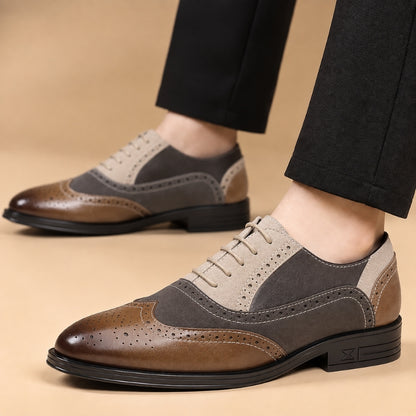 A pair of men's wingtip oxford shoes in brown, gray, and beige suede and leather, shown on a model wearing black trousers against a warm beige studio background.