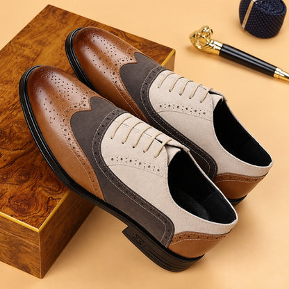 A pair of men's wingtip oxford shoes in brown and beige suede-like material with dark gray panels, displayed on a wooden box against a beige background with a pen and pocket square prop.