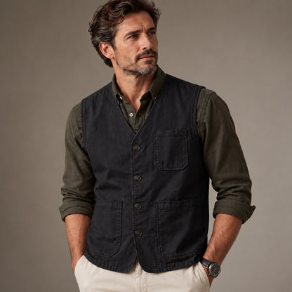 A man wearing a dark charcoal gray utility vest over an olive green button-down shirt and cream pants, standing against a neutral gray studio background.
