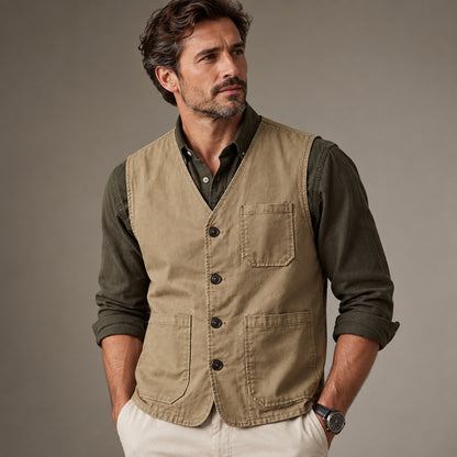 A man wearing a tan utility vest over an olive green button-down shirt and cream pants, standing against a neutral gray studio background.