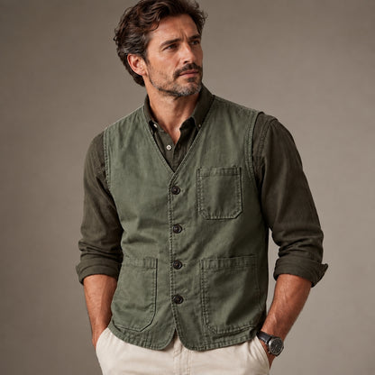 A man wearing an olive green button-front vest over a dark long-sleeve shirt and off-white pants, standing against a plain taupe studio background.