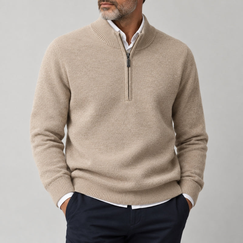 A man wearing a light beige half-zip knit sweater over a white collared shirt and navy trousers, shown from the chin down against a plain light gray studio background.