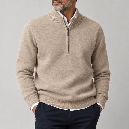 A man wearing a light beige half-zip knit sweater over a white collared shirt and navy trousers, shown from the chin down against a plain light gray studio background.