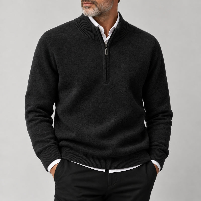 A man wearing a dark charcoal gray half-zip sweater over a white collared shirt and black trousers, shown from the chin down against a light gray studio background.