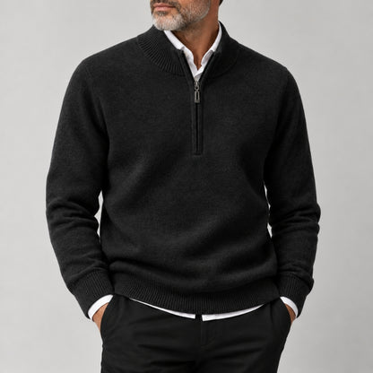 A man wearing a dark charcoal gray half-zip sweater over a white collared shirt and black trousers, shown from the chin down against a light gray studio background.