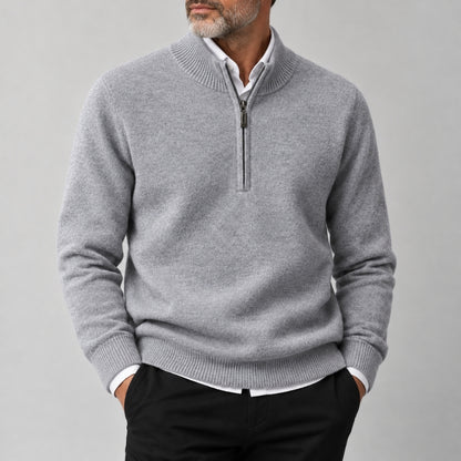 A man wearing a light gray half-zip knit sweater over a white collared shirt and black trousers, standing against a plain light gray studio background.
