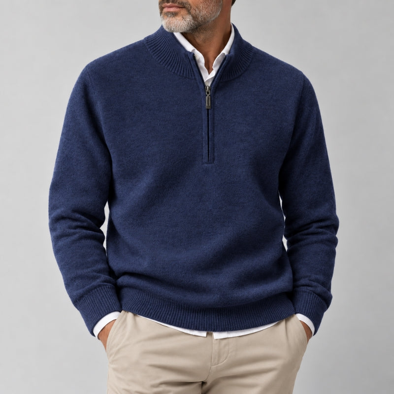 A man wearing a navy blue half-zip sweater over a white collared shirt and beige trousers, shown from the chin down against a plain light gray studio background.