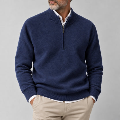 A man wearing a navy blue half-zip sweater over a white collared shirt and beige trousers, shown from the chin down against a plain light gray studio background.