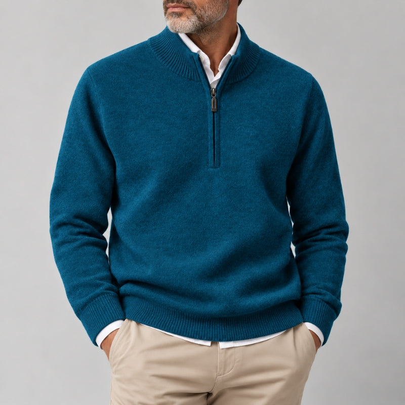 A man wearing a teal blue quarter-zip knit sweater over a white collared shirt and beige trousers, shown from the chin down against a plain light gray studio background.