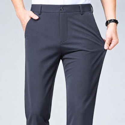 A close-up of a person wearing dark charcoal gray dress pants, showing the front waistband, front pockets, and side pockets being stretched by the hand. The person is cropped from the waist down, with a light gray studio background.