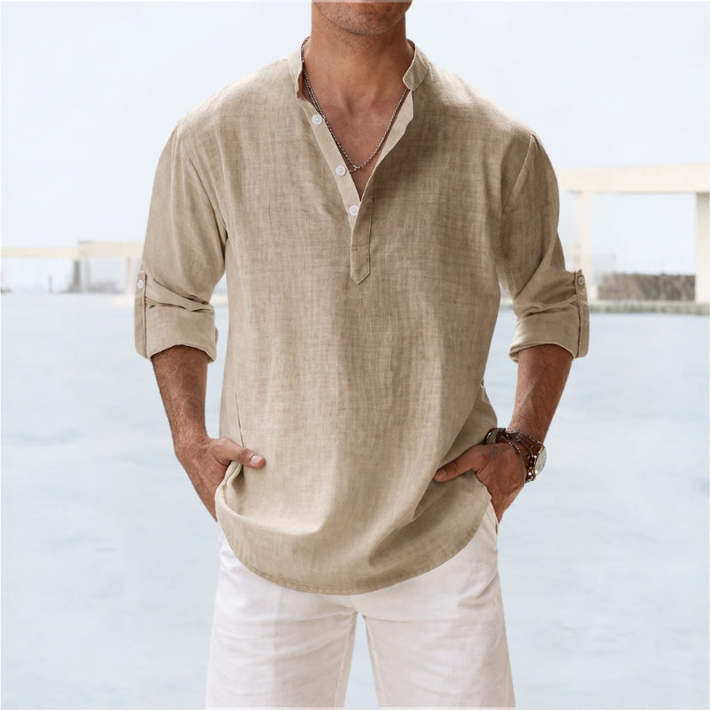 Man wearing a khaki linen shirt with rolled-up sleeves and white pants against a light background