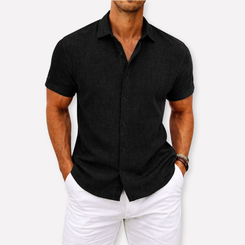 Man wearing a black short-sleeve button-up shirt and white pants against a white background