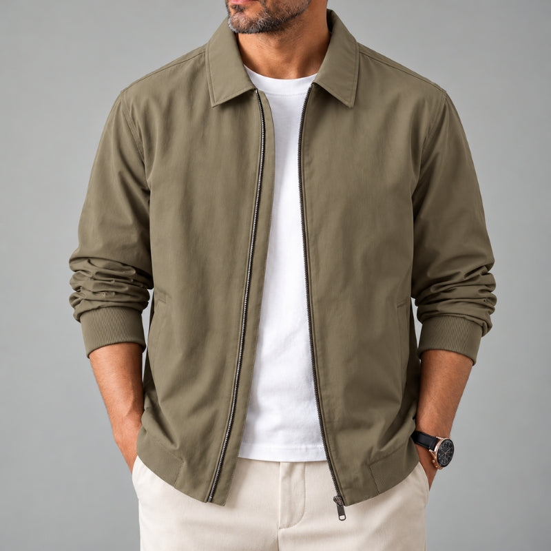 A man wearing an olive green zip-up jacket over a white t-shirt and beige pants, shown from the chin down against a solid gray background.
