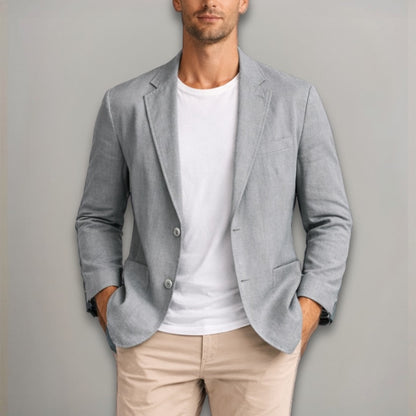 Man wearing a light gray blazer over a white shirt and beige pants against a plain background