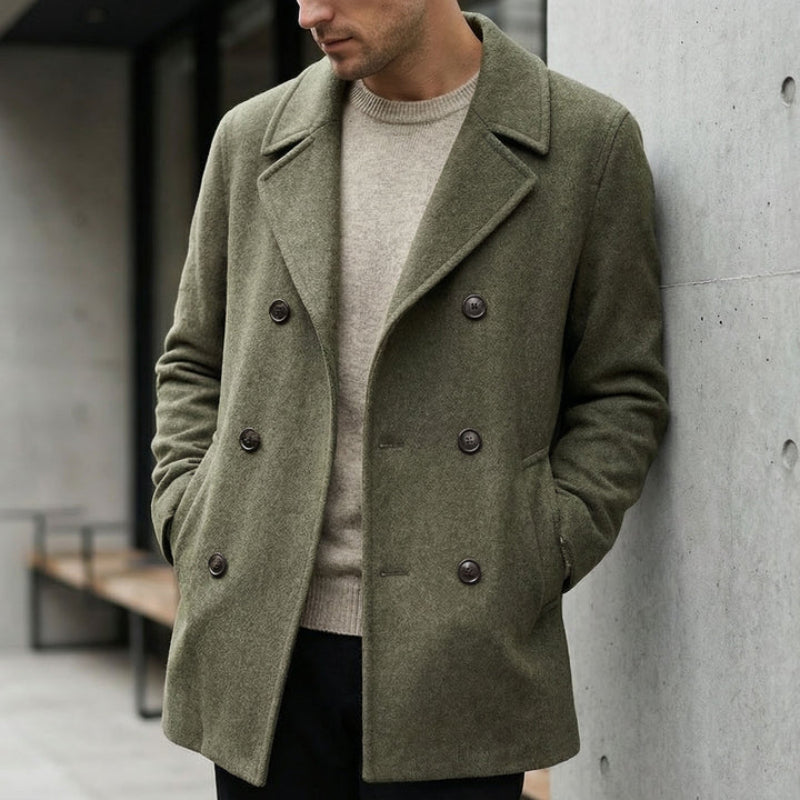 Man wearing a green coat leaning against a concrete wall.