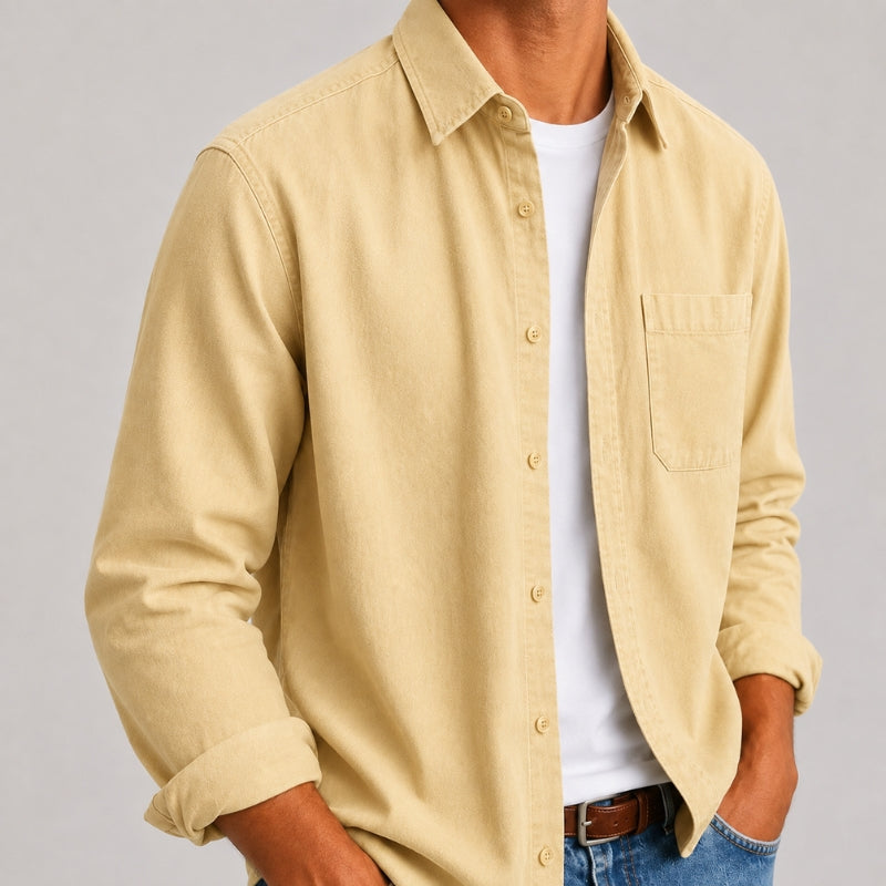 A man wearing a light beige long-sleeve button-down shirt over a white t-shirt and blue jeans, shown from the chest down against a neutral gray background.