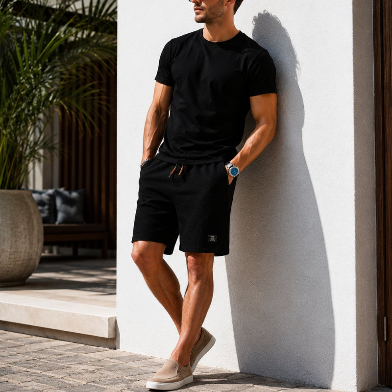A man wearing a black short-sleeve t-shirt and black drawstring shorts leans against a white wall in a modern outdoor setting. He is wearing beige slip-on shoes and a blue watch.