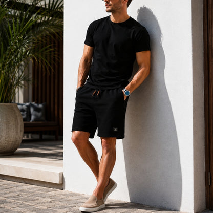 A man wearing a black short-sleeve t-shirt and black drawstring shorts leans against a white wall in a modern outdoor setting. He is wearing beige slip-on shoes and a blue watch.