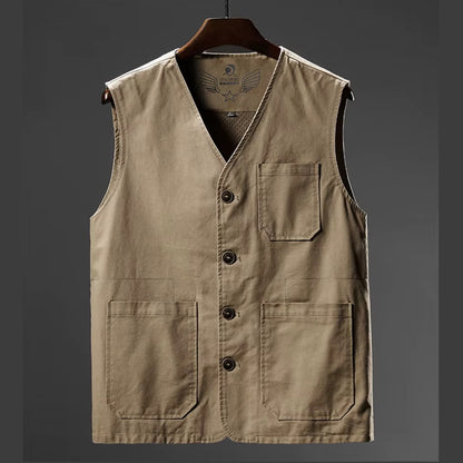 A sleeveless utility vest in a warm khaki-beige color, hanging on a dark wooden hanger against a dark gray gradient background. The vest features a V-neck, a button-up front, a chest pocket, and two large front patch pockets.